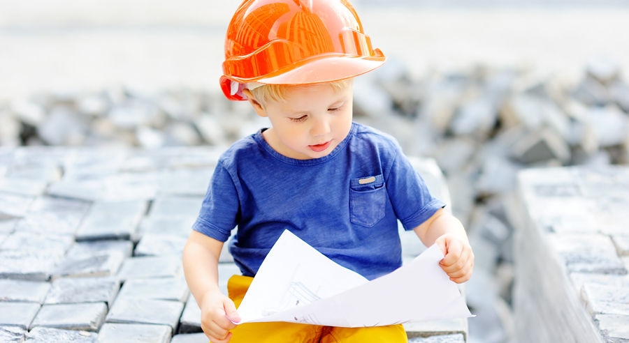 Kleiner Junge sitzt mit Helm auf einer Baustelle und sieht sich den Plan an