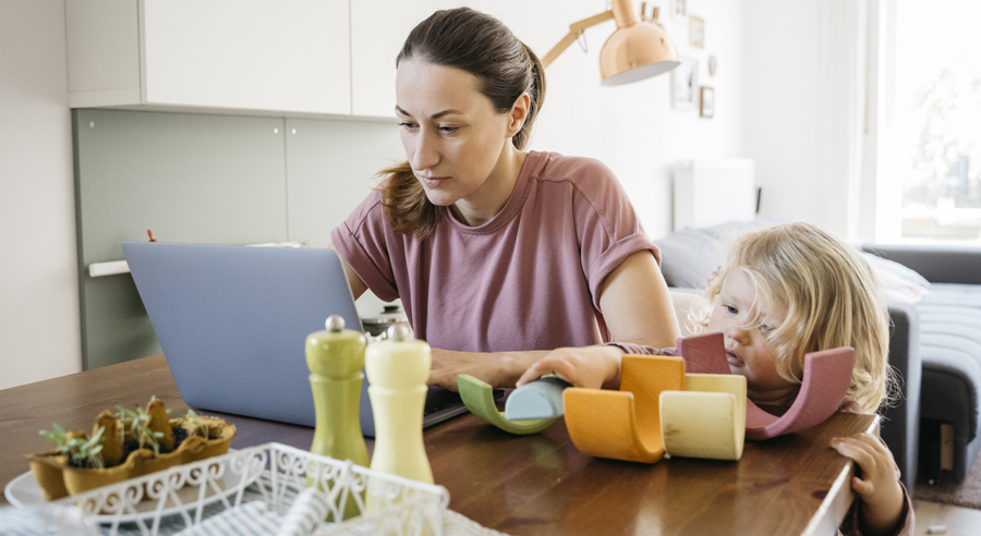 Eine Frau sitzt mit ihrer kleinen Tochter am Tisch und arbeitet im Homeoffice am Laptop