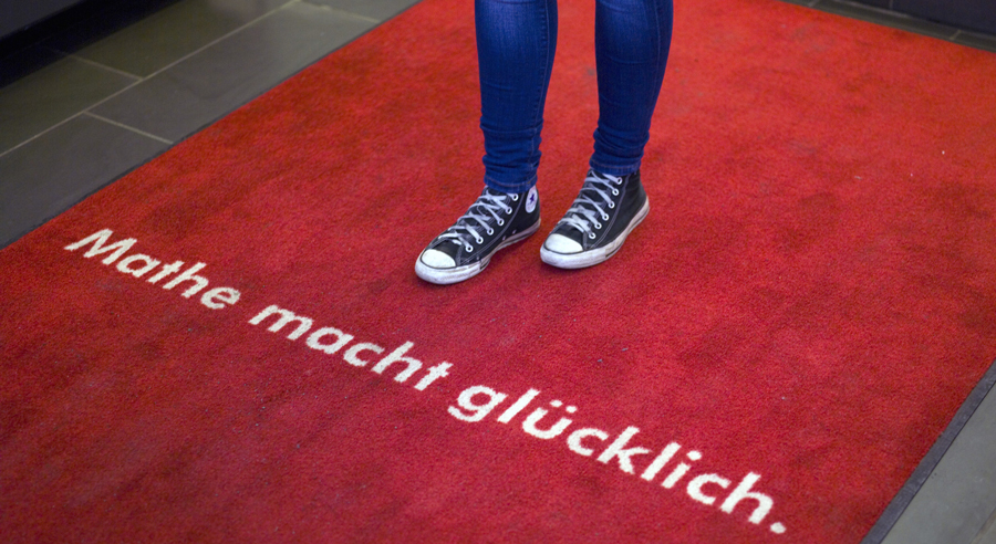 Die Beine eines Mädchens in Jeans stehen auf einem roten Teppich mit der Aufschrift „Mathe macht glücklich“