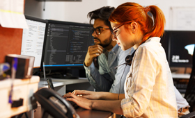 Eine junge Frau und ein junger Mann sitzen an einem Schreibtisch vor einem PC und programmieren